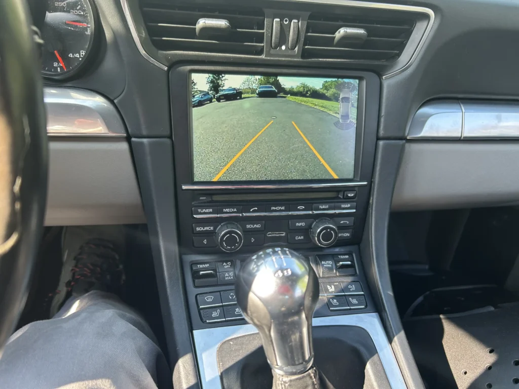 Porsche center console displaying a backup camera feed with yellow trajectory guide lines and parking sensors, installed by Skylands Car Audio and Accessories.
